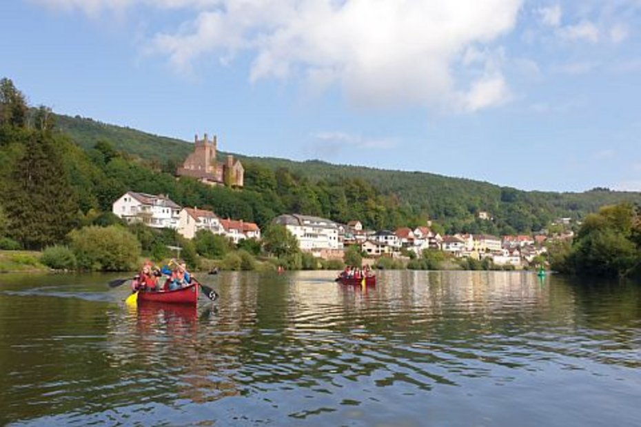 Mehrere Personen paddeln in Kanus auf einem ruhigen Fluss, im Hintergrund sind Häuser am Ufer und eine Burg auf einem bewaldeten Hügel unter blauem Himmel zu sehen.