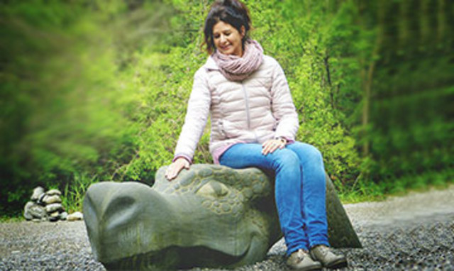 Vor einem grün belaubten Hintergrund liegt die Skulptur eines steinernen Drachenkopfes. Auf diesem sitzt eine Frau mittleren Alters und streichelt dem Drachen über den Kopf.