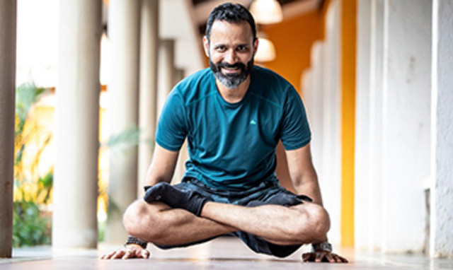 Ein Mann mit dunklen, leicht grau melierten Haaren und Bart macht eine Yogaübung im Schneidersitz und stemmt sich dabei vom Boden ab. Er lächelt freundlich in die Kamera.