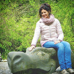 Vor einem grün belaubten Hintergrund liegt die Skulptur eines steinernen Drachenkopfes. Auf diesem sitzt eine Frau mittleren Alters und streichelt dem Drachen über den Kopf.