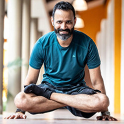Ein Mann mit dunklen, leicht grau melierten Haaren und Bart macht eine Yogaübung im Schneidersitz und stemmt sich dabei vom Boden ab. Er lächelt freundlich in die Kamera.